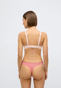 Gaiši rozā krūšturis ar regulējamām lencēm un gludu faktūru, kopā ar rozā tanga biksītēm. Skats no aizmugures atklāj minimālistisku dizainu un bezšuvju malas.