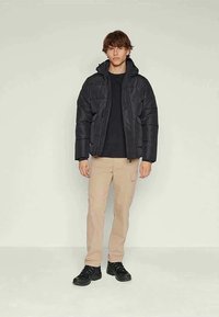 Chaqueta negra inflada con capucha y cierre de cremallera, fabricada en una mezcla de algodón, combinada con pantalones cargo beige y zapatillas negras.