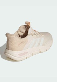 Zapatilla de correr beige claro con parte superior de malla texturizada, rayas blancas y suela acolchada. Cuenta con una lengüeta para tirar y un diseño con branding minimalista.