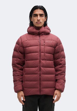 Hombre con cabello negro que lleva una chaqueta acolchada burdeos con un cuello alto, de pie frente a un fondo liso gris claro.