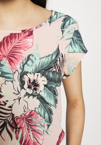 Haut à motif floral en tissu doux et texturé. Présente un fond rose avec des feuilles et des fleurs vibrantes dans des teintes de vert et de rouge. Encolure ronde.