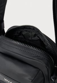Sort nylon crossover taske med lynlås åbning, der har en glat overflade og et ekstra rum indeni, med subtil logo detalje synlig.
