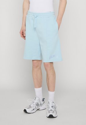 Person som har på seg lyseblå shorts med snøring og teksten "Colucci", hvite sokker og hvite New Balance-sneakers, står mot en ensfarget bakgrunn.