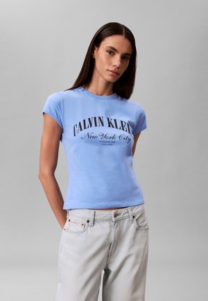 Jeune femme portant un t-shirt bleu clair Calvin Klein New York City et un jean gris clair, debout devant un fond gris uni.
