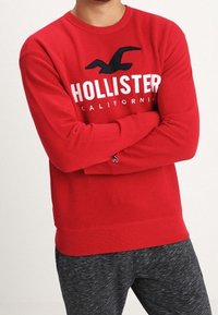 Sweatshirt rouge à manches longues arborant le texte "HOLLISTER CALIFORNIA", un logo d'oiseau, des poignets côtelés et un tissu lisse. Associé avec un bas gris foncé.