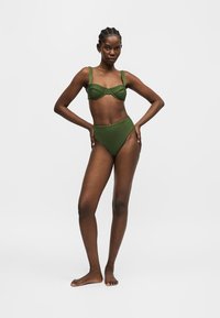 Femme debout pieds nus portant un soutien-gorge à armatures vert olive et des culottes taille haute sur un fond clair uni.