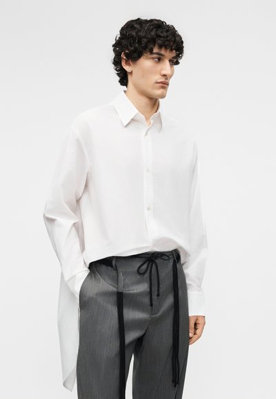 Junger Mann mit lockigen Haaren, der ein locker sitzendes weißes Hemd mit Knöpfen und graue Nadelstreifenhosen mit schwarzen Kordeln trägt, Hände in den Taschen, schaut nach rechts.