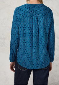 Blouse à manches longues en bleu vif avec un motif en losange, tissu texturé, ourlet arrondi et poignets boutonnés.