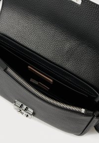 Μαύρη τσάντα ώμου από ανάγλυφο δέρμα με ασημένιο κούμπωμα, ανοιχτή για να αποκαλύψει ένα φερμουάρ τμήμα και εσωτερικές θήκες.