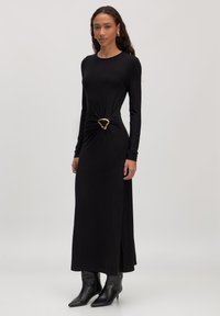 Zwarte lange mouwen jurk met een knoopdetail in de taille en een gouden hartvormige gesp. Soepele stof, enkel-lengte snit, gedragen met spitse zwarte laarzen.