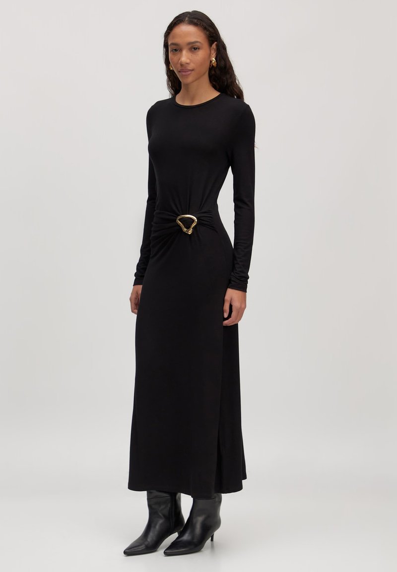 Zwarte lange mouwen jurk met een knoopdetail in de taille en een gouden hartvormige gesp. Soepele stof, enkel-lengte snit, gedragen met spitse zwarte laarzen.