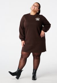 Bruine jurk met lange mouwen en een losse pasvorm, voorzien van een ronde halslijn en subtiele zijslits. Gecombineerd met zwarte enkellaarsjes en doorschijnende panty's.