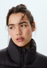 Čierna prešívaná bunda s vysokým golierom a detailmi na zips, s hladkou, lesklou textúrou. Detailný záber na tvár s jemným make-upom a rozcuchanými vlasmi.