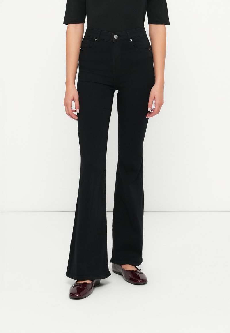 Personne portant un pantalon évasé noir taille haute, un haut noir, et des ballerines en cuir verni rouge foncé, se tenant contre un fond blanc.