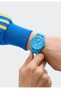 Sinine plastist kell, millel on ümmargune nägu ja minimalistlik disain. Rihmal on tekstuuriga viimistlus ja reljeefne logo.