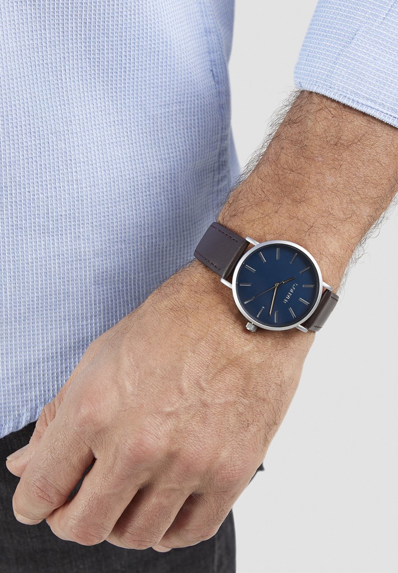 Montre-bracelet argentée avec un cadran rond bleu, un bracelet en cuir brun et des accents argentés. La montre présente des marqueurs d'heure simples et une couronne.