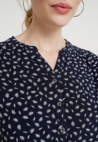 Mörkblå knappad blus med vitt bladmönster, V-ringning, mjukt tyg och sammetsärmar för extra textur och detalj.