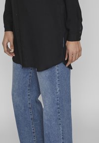 Chemise noire à manches longues avec des boutons et des fentes sur les côtés, associée à un jean large bleu clair, présentant une texture en denim doux et des coutures visibles.