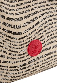Tkaninska torba z bež ozadjem in ponovljenim črnim napisom "JOOP!JEANS" ter rdečim embosiranim okroglim logotipom.