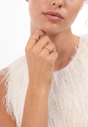 Deux fines bagues en or avec de petites pierres précieuses sont portées aux doigts d'une main reposant près du visage ; en arrière-plan, un vêtement blanc texturé.