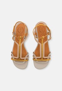 Beige nahka sandaalit, joissa on kultaiset metalliset hihnat, avoin varvasmuotoilu ja tasapohja. Mukana säädettävä nilkkahihna.