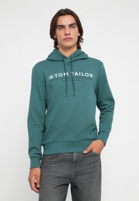 Felpa teal con tasca a canguro frontale, cordini regolabili e testo del logo bianco. Abbinata a jeans grigi scuri.