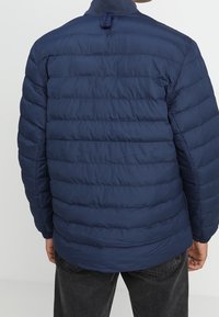 Marinblå puffjacka med quiltad design och hög krage. Utrustad med en ögla vid nacken och elastiska muddarna för en tät passform.