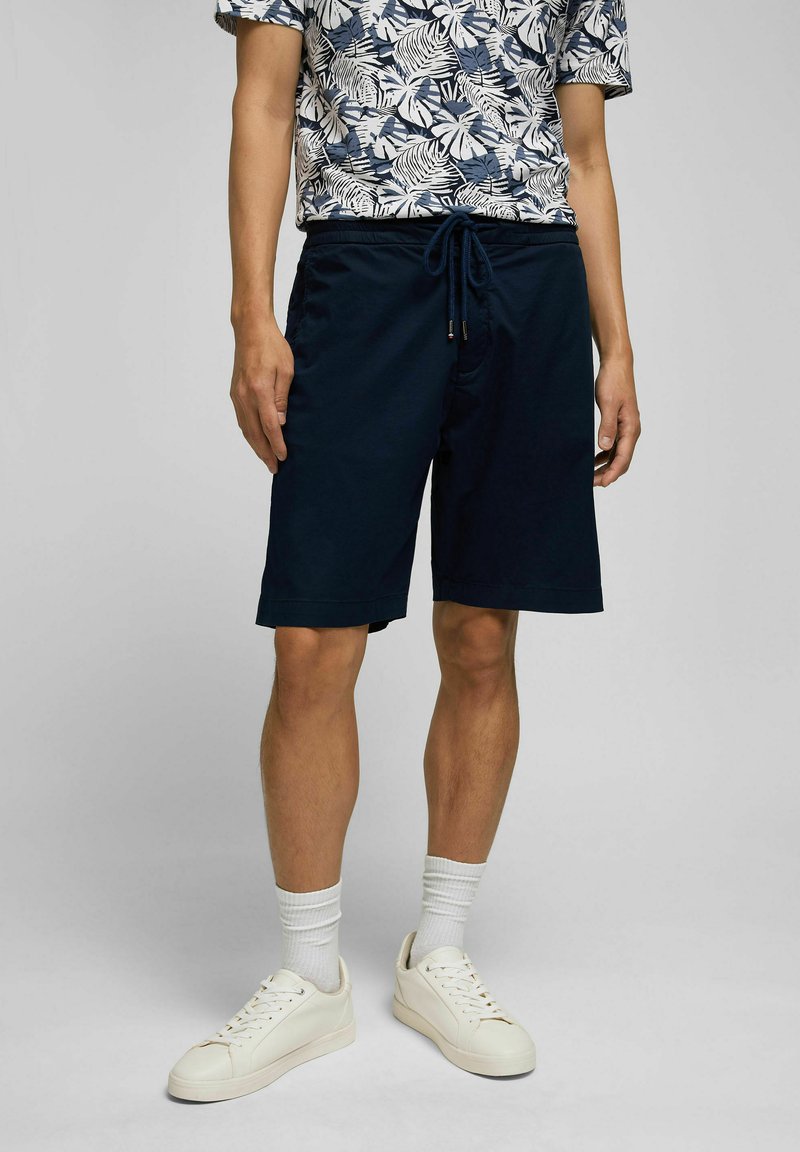 Marineblaue Shorts mit elastischem Bund, Kordelzug und Seitentaschen. Getragen mit einem gemusterten Hemd und weißen Sneakern.