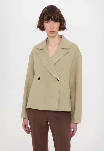 Veste en mélange de laine beige avec un design croisé, des revers à cran, deux poches et des boutons noirs. Dispose de manches oversized légèrement raccourcies.
