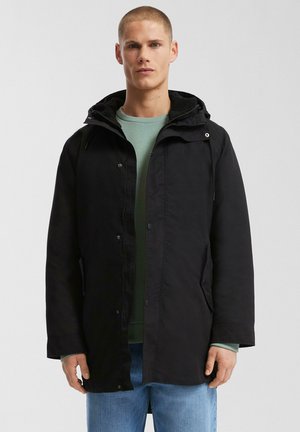 Schwarze Parka mit Kapuze, ausgestattet mit einem Reißverschluss und Druckknopfverschluss, Seitentaschen und einem weichen Futter. Getragen über einem hellgrünen Hemd.