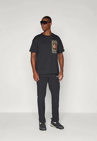 Vyras, vilkintis juodą marškinėlį su grafika, juodas kelnes, juodus sportinius batus ir didelius juodus akinius nuo saulės, stovi prieš paprastą šviesią foną.