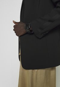 Osoba s tmavou pletí v černém saku a béžové saténové sukni, nosící černé kovové hodinky na levém zápěstí, s rukou umístěnou poblíž saka.