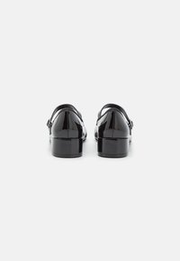 Black patent leather shoes feature a low block heel, ankle strap, and rounded toe. The shiny texture enhances visual appeal.