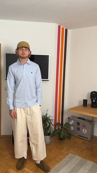 Camisa azul claro, com botões e mangas longas, e um pequeno logo, combinada com calças largas de cor creme e tamancos castanhos.