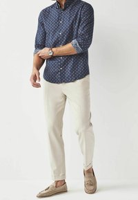 Man die de mouwen oprolt van een marineblauwe gestreepte overhemden, draag beige broek en beige loafers met kwastjes, op een witte achtergrond.