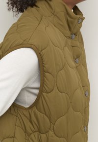 Chaleco acolchado verde oliva con botones a presión, cuello alto y diseño sin mangas. Textura suave con patrones acolchados circulares y ribete contrastante.