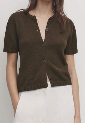 Femme portant un cardigan à manches courtes, boutonné, de couleur marron foncé, associé à un pantalon taille haute blanc cassé, debout les mains dans les poches.