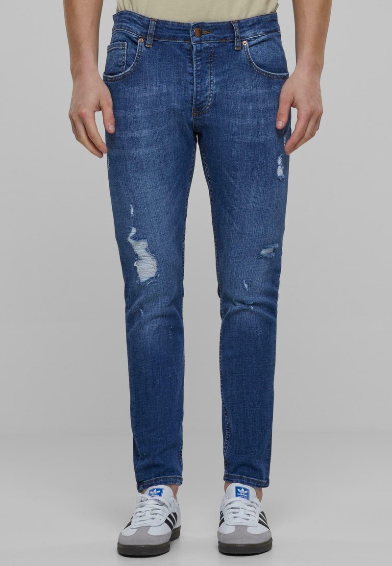 Megkopott kék farmernadrág, slim fit szabással, elhasználódott részletekkel és gombos záródással. Fehér és szürke sportcipőkkel párosítva.