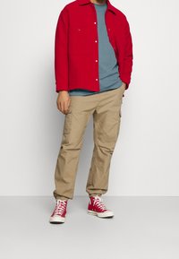 Uomo che indossa una giacca rossa con bottoni, una maglietta blu, pantaloni cargo beige e scarpe da tennis alte rosse, in piedi su uno sfondo bianco semplice.