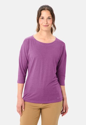 Frau mit zurückgebundenen braunen Haaren, die ein lila Dreiviertelarm-Oberteil und beige Hosen trägt, steht vor einem schlichten Hintergrund.