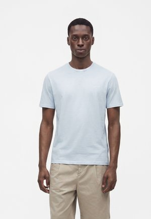 Joven con el pelo corto que lleva una camiseta de manga corta azul clara y pantalones beige, de pie frente a un fondo blanco.