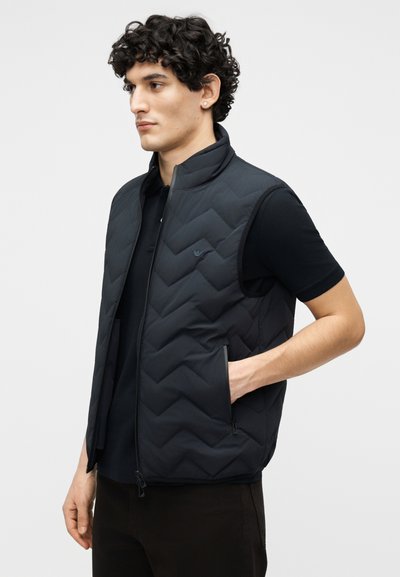 Άνδρας που φοράει μαύρο αμάνικο μπουφάν με ραφές πάνω από μαύρο κοντομάνικο πουκάμισο, στέκεται με το ένα χέρι στην τσέπη του μπουφάν, κοιτώντας προς τα αριστερά.