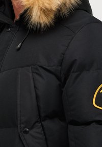 Veste matelassée noire avec une capuche bordée de fourrure. Elle présente un tissu texturé, des coutures visibles, et un logo circulaire jaune sur la manche.
