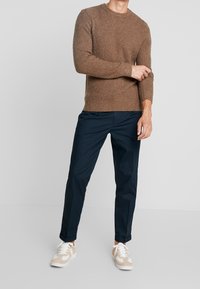 Man justerar ärmen på en brun texturerad tröja, bärande marinblå byxor och beigea och vita sneakers, stående mot en enkel ljus bakgrund.