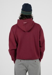 Bordowy sweatshirt z kapturem, z kieszenią typu kangur i ściągaczami na rękawach, z haftem na plecach. W połączeniu z szarymi wełnianymi spodniami i granatową czapką.