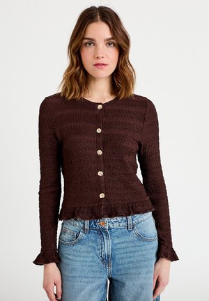 Vrouw met schouderlang bruin haar, gekleed in een getextureerde donkerbruine blouse met knopen en een lange mouw, en lichtblauwe jeans met een hoge taille.