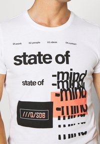 Bílý bavlněný tričko s výrazným černým textem, oranžovými akcenty a grafickými vzory. Text zahrnuje fráze jako "stav mysli" a "kontakt".