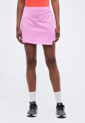 Femme portant une jupe de sport rose, des chaussettes blanches, des chaussures de sport noires et un haut orange, debout devant un fond blanc uni.