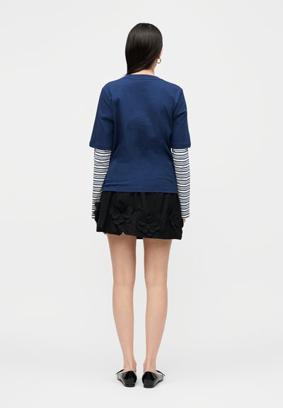 Γυναίκα που φοράει μπλε σκούρο κοντομάνικο τοπ πάνω από μακρυμάνικο λευκό και μπλε ριγέ πουκάμισο, μαύρη φλοράλ φούστα και μαύρα παντόφλες, από πίσω.