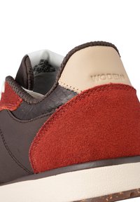 Athletic shoe featuring brown leather, red suede accents, and a beige leather heel tab, with textured surfaces and stitched seams.